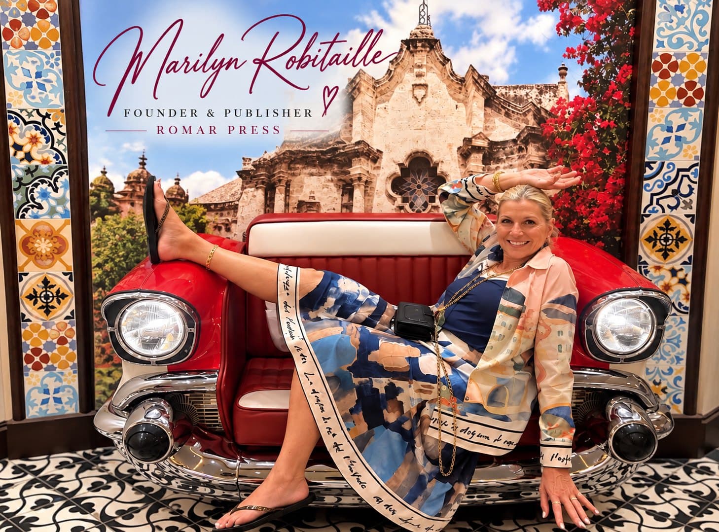 Dr. Marilyn Robitaille, Founder and Publisher of ROMAR Press, smiling on a red vintage-car bench in front of a painted cathedral, framed by patterned tiles