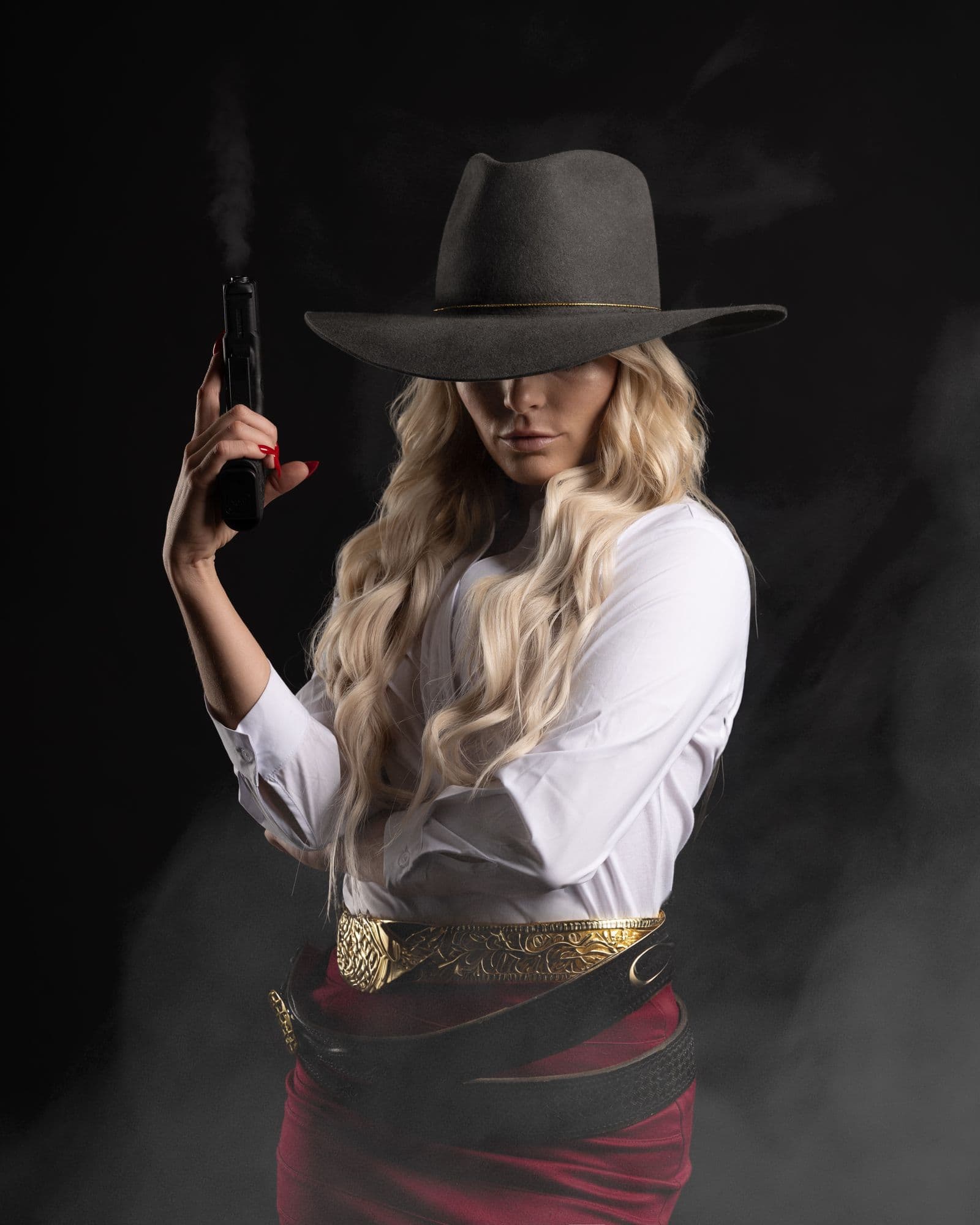 A woman in a dark cowboy hat, white shirt and red skirt raises a pistol through drifting smoke